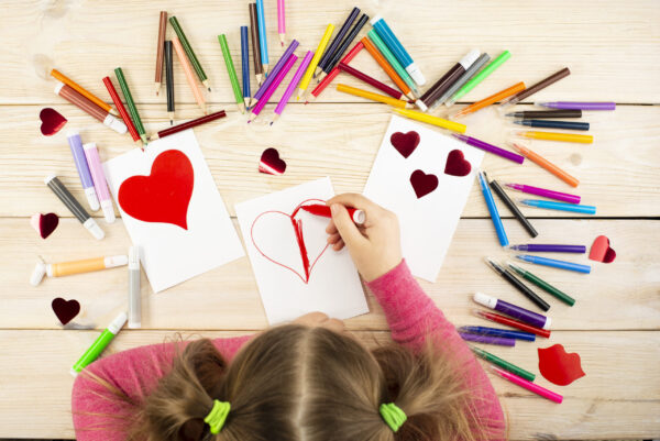Little girl prepares cards with hearts for Valentine's Day. Drawing and applique work is done by the child using colored pencils or felt-tip pens. Children's drawing. View from above