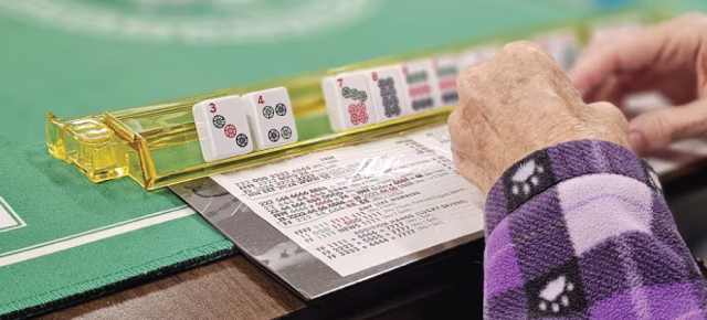 Close-up of hands arranging Mahjong tiles on a green table, accompanied by a Mahjong card and tile rack.