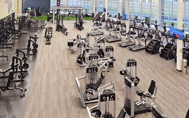 A spacious and modern gym with a variety of exercise equipment, including treadmills, stationary bikes, weight machines, and free weights. The gym features large windows along one side, providing natural light and a view of the outdoors.
