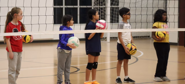 boys and girls wait for instruction behind a volleyball net while holding volleyballs