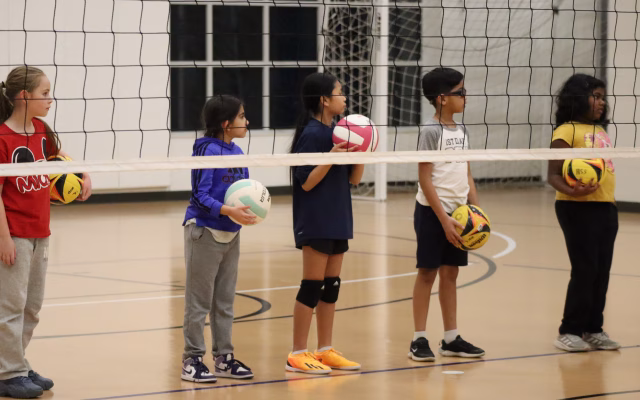 boys and girls wait for instruction behind a volleyball net while holding volleyballs