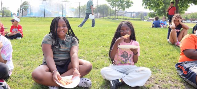 Two girls sitting in grass eating pizza