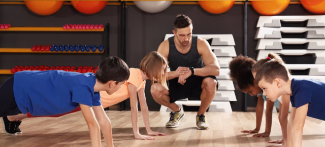 children in a push up position while a coach is kneeled next to the looking at his watch