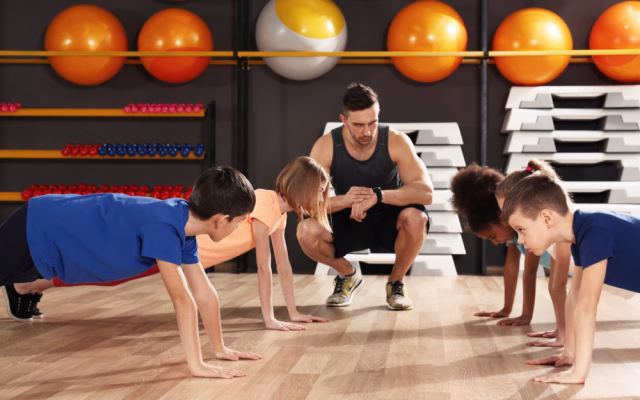 children in a push up position while a coach is kneeled next to the looking at his watch