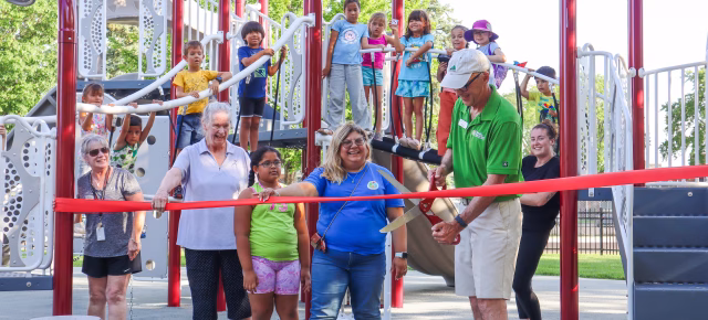 Group of adults and children at a playground ribbon-cutting ceremony, with a man in a green shirt cutting a red ribbon while smiling. Children are standing on playground equipment in the background, and everyone appears happy and excited.
